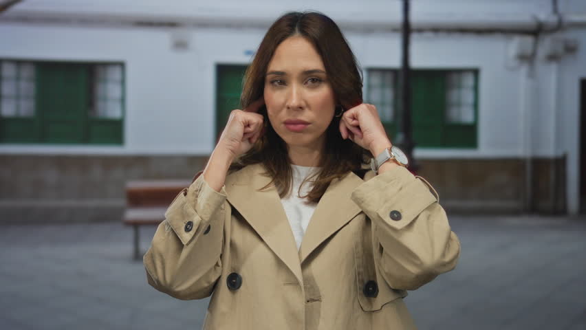 Young woman in beige coat standing on city street, covering ears with fingers, expressing annoyance or frustration in urban outdoor setting with white building background.