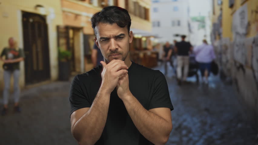 Young hispanic man appearing thoughtful on a busy city street in an outdoor urban setting, surrounded by blurred pedestrians and rustic buildings.