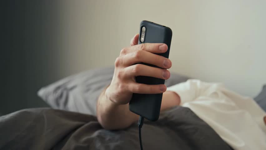 Side stab shot of a person unplugging a charger from a smartphone while lying in bed in the morning at home, symbolizing daily routine, technology use, and modern lifestyle.