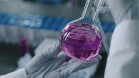 Close-up shot of the hands of an anonymous female scientist in a white coat and gloves examining a glass flask with a light purple chemical solution during laboratory research. - Powered by Shutterstock - Get 15% off with code: PIKWIZARD15