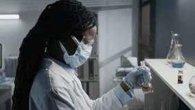 Side shot of a focused female Black medical researcher wearing goggles, mask, and gloves, adding reagent to a solution in a flask while developing a new revolutionary drug. - Powered by Shutterstock - Get 15% off with code: PIKWIZARD15