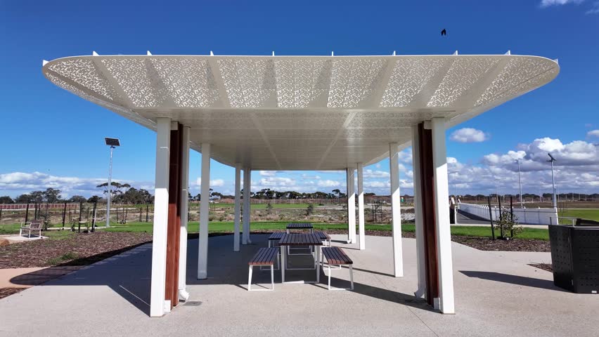 Modern outdoor shelter with picnic tables in an Australian suburban park, featuring decorative roof design and open community space. Public recreation, urban lifestyle, and family-friendly facility