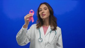 Hispanic woman doctor with stethoscope holds pink ribbon against blue background, symbolizing breast cancer awareness and care. - Powered by Shutterstock - Get 15% off with code: PIKWIZARD15