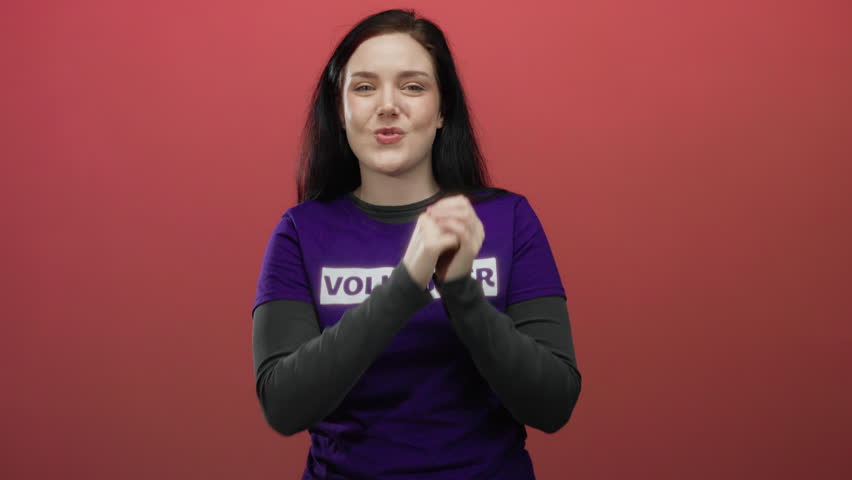 Beautiful brunette woman wearing volunteer shirt claps joyfully in front of vibrant red background radiating happiness and engagement