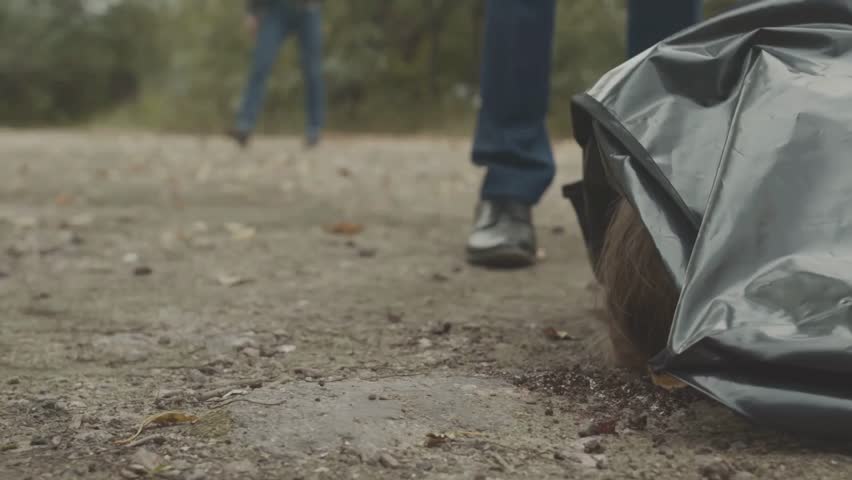 A tense moment unfolds as two detectives stand over a covered body at a city park crime scene.
