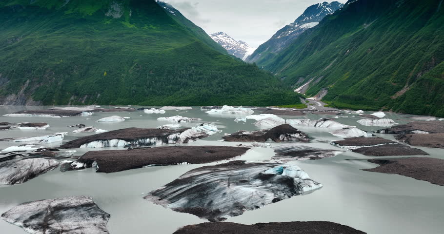 Ice lumps sticking out of grey riverscape. Huge verdant mountains on the waterfront at backdrop. Alaska, USA.