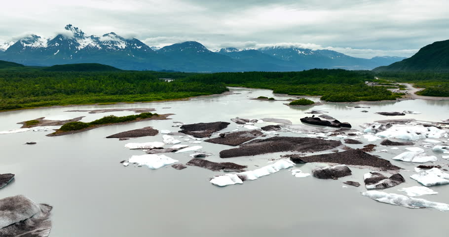 Flying over the river with melting ice approaching the green waterfront. Snow-capped mountains covered with clouds at backdrop. Alaska, USA.