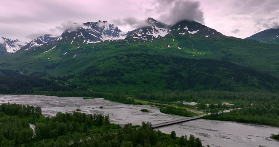 Flight over the river crossed by the automobile bridge. Grey clouds cover the snow-capped tops of the mountains. Nature of Alaska, USA.