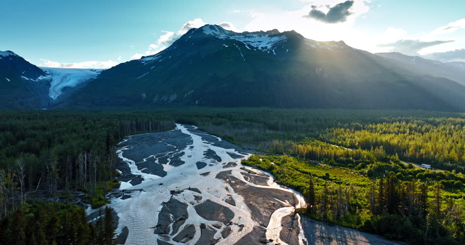 Shallow branching river in the valley covered with pine tree woods. Sun hides behind the snow-capped rock. Alaska, US. Aerial view.