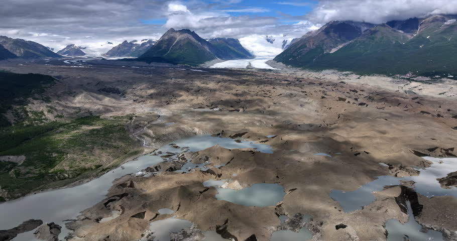 Uneven landscape with some water. Clouds throw shadows on the ground. Grey fluffy clouds cover the snow-capped rocks. Alaska wilderness. Aerial view.