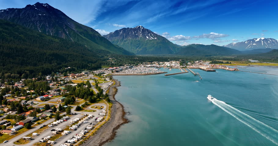 Little town located on the waterfront of the river in the mountains. Cruise boat goes towards the shore. Alaska, USA. Aerial view.