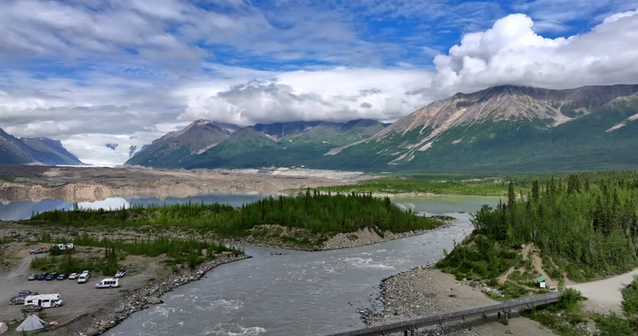 Footage over the rapid turbulent river flowing at the mountain foot. Beautiful white cloudscape cover the mountain peaks at backdrop. Alaska, USA.