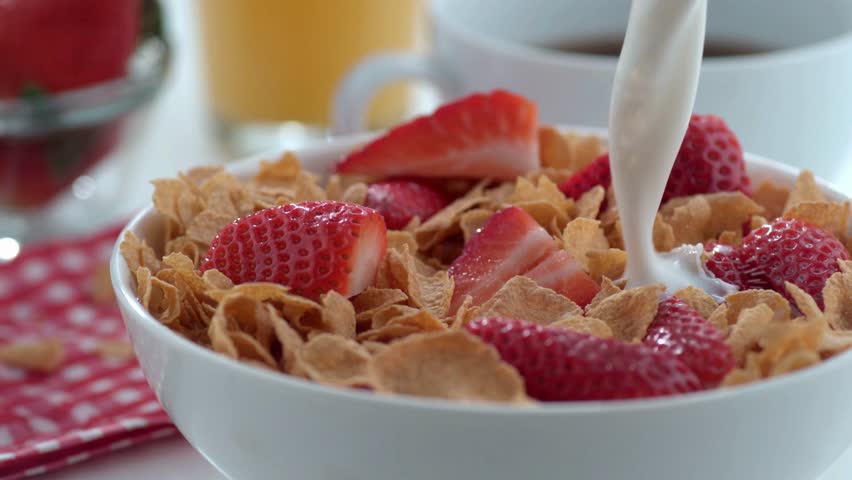 Close-up slow-motion shot of milk being poured over a bowl of breakfast cereal and fresh strawberries, creating a classic, refreshing morning scene. Advertising style.