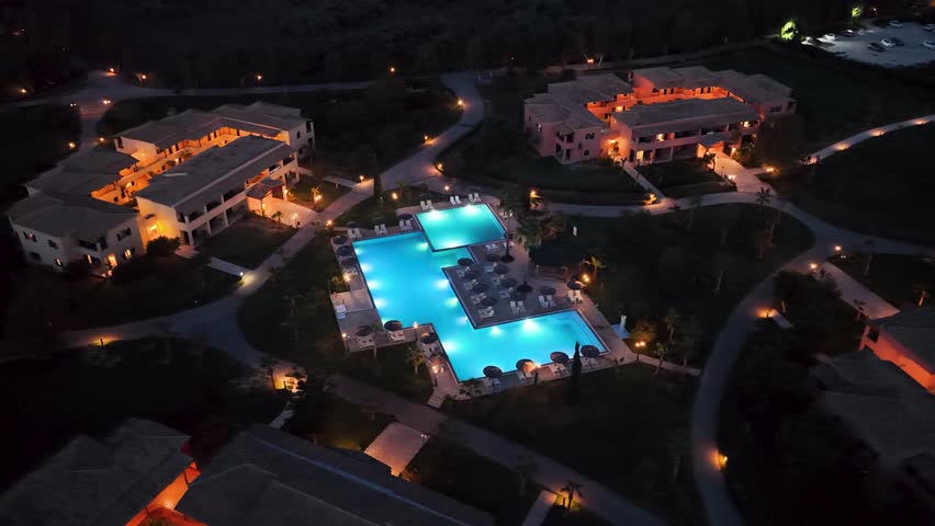 Aerial drone view of a large, luxury resort swimming pool illuminated with bright blue lights at night.