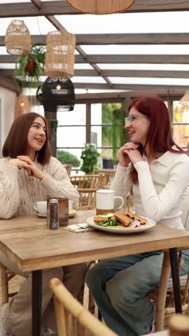 Vertical screen two women sit in a cozy cafe smiling warmly at each other as they share sweet compliments and supportive words creating a gentle uplifting atmosphere during a relaxed breakfast 