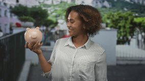 Woman holding piggy bank on street, smiling with hands visible and striped shirt; saving money confidence joy. - Powered by Shutterstock - Get 15% off with code: PIKWIZARD15