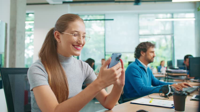 Smiling Caucasian woman using smartphone sitting at desk during workday. Female employee scrolling social media while colleagues focused on tasks nearby. Relaxed moment in shared office space.