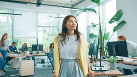 Confident Asian woman standing with arms crossed and smiling slightly to camera. Female professional in casual outfit posing in bright office while coworkers working behind at desks. - Powered by Shutterstock - Get 15% off with code: PIKWIZARD15
