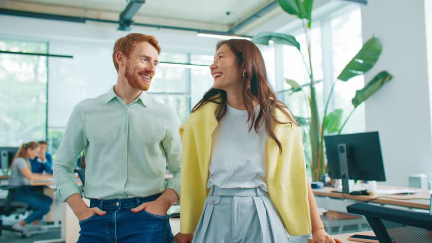 Two colleagues from work standing side by side with arms crossed while smiling brightly. Open workspace with computers and plants in blurred background. Showing team cooperation and confidence.