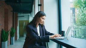 Charming Asian businesswoman sitting by window looking confidently at camera. Wearing dark blazer and white top. Laptop and coffee cup on desk. Greenery outside visible behind glass window. - Powered by Shutterstock - Get 15% off with code: PIKWIZARD15