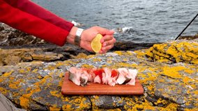 A close-up of a person seasoning fresh fish slices with lemon juice and olive oil outdoors by the seaside. The fish is placed on a wooden board over colorful coastal rocks, with sea waves. - Powered by Shutterstock - Get 15% off with code: PIKWIZARD15