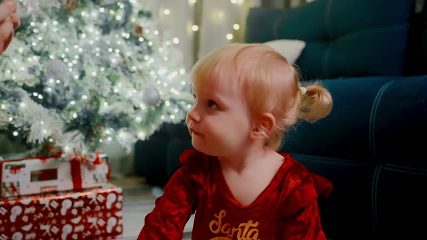 mother and toddler prepare holiday accessories, young child with holiday adornment near christmas tree, child gently dressed with seasonal headband next to decorated tree