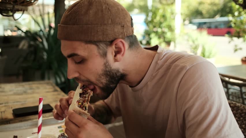 A young man takes a bite of a burrito while sitting in an outdoor cafe on a sunny day. Guy eating Mexican fast food. Horizontal 4k footage
