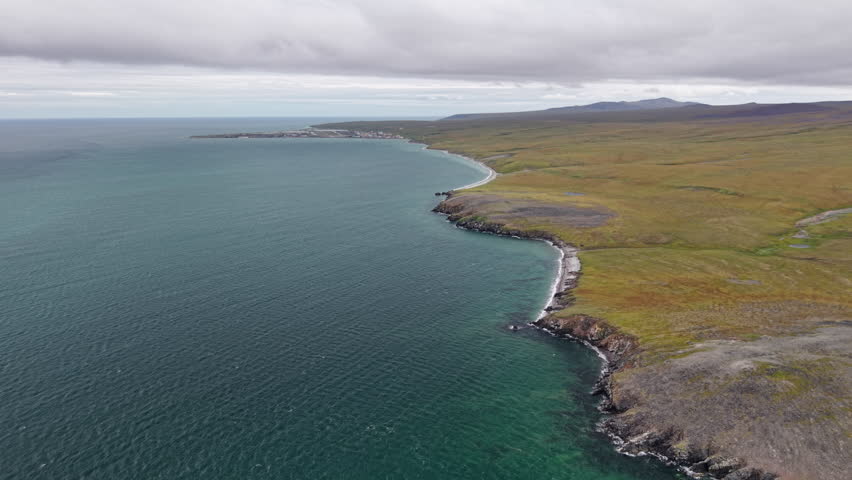 Endless tundra coast stretches along Lavrentiya Bay in Chukotka with a village on the horizon. Gentle surf and open space convey remoteness and calm ideal for travel, tourism and documentary use
