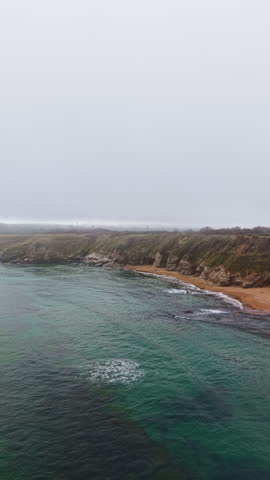 Misty morning unfolds over a tranquil beach as waves kiss the sandy shore. The rugged cliffs rise majestically, and the calm sea mirrors the muted colors of dawn.