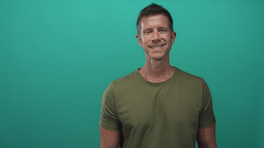 Man extending right hand for handshake and smiling in a teal studio with olive t shirt and bracelets; friendly welcome.