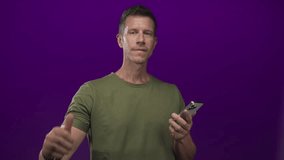 Man giving thumbs up while holding smartphone and displaying bare hand in purple studio setting, wearing olive t shirt; approval confidence. - Powered by Shutterstock - Get 15% off with code: PIKWIZARD15