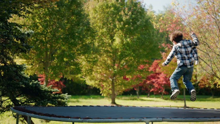 Little boy bouncing on trampoline in sunny park surrounded by green and colorful autumn trees. High quality 4k footage