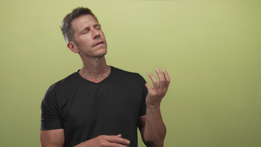 Man examining and pointing at his fingers while wearing black tshirt in studio with green wall; thoughtful.