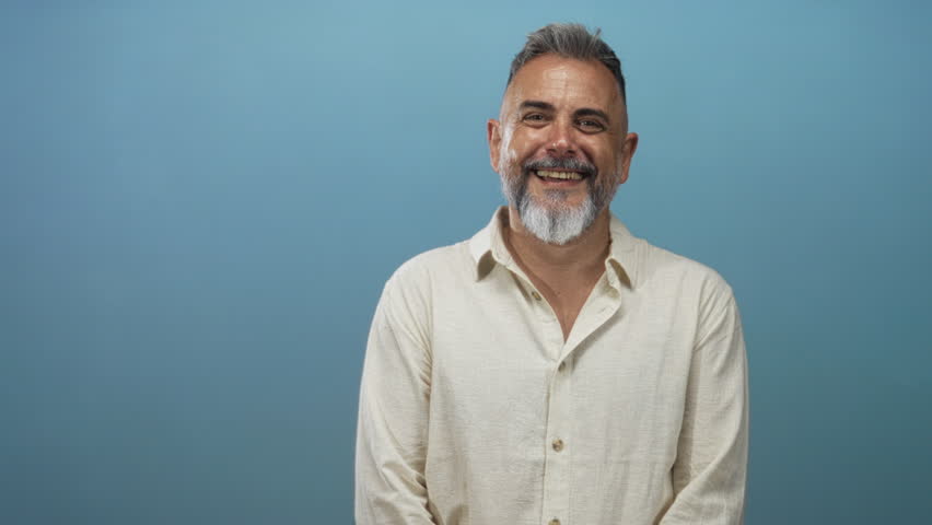 Middle aged man smiling and raising hand, bearded and wearing linen shirt in blue studio, relaxed posture and open smile; joy confidence.