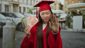 Woman in red graduation gown holds philippines peso banknotes and gestures stop on urban street background indicating financial caution outdoors. - Powered by Shutterstock - Get 15% off with code: PIKWIZARD15