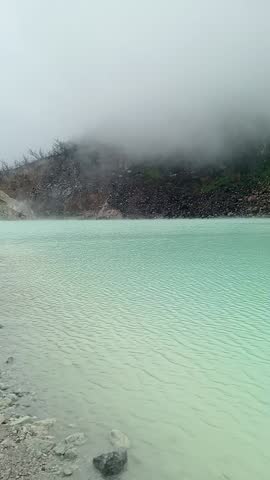 A misty and foggy light green lake in a white crater with a hill in the background and several people seen on a wooden bridge.