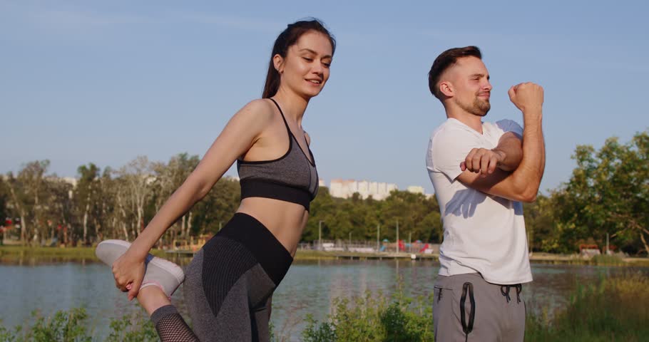 Sporty happy fit couple outdoor, woman moving her arms, man leg stretching and warming up, friends exercising together, promoting health and fitness joy, enjoying fresh air near river, lake fun