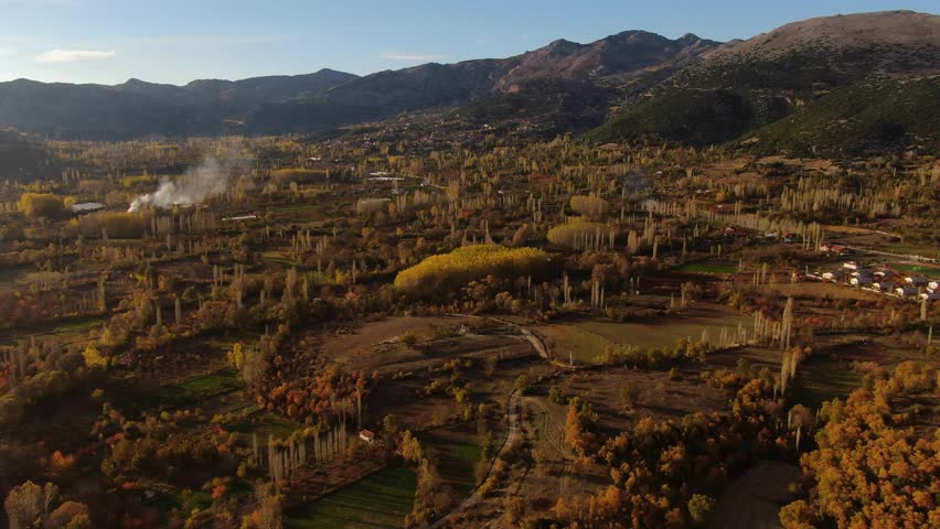 A settlement located in the middle of nature, in a valley filled with farmland and deciduous trees. Smoke rises in the distance.