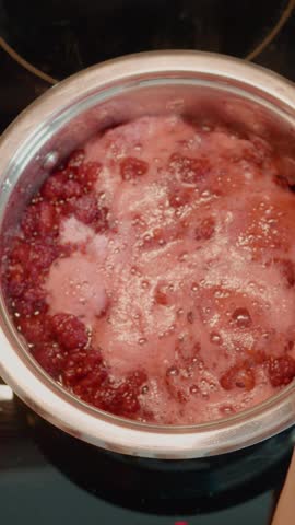 Vertical Screen: In a cozy kitchen, a woman stirs boiling raspberries in a saucepan, creating a delicious homemade jam while enjoying the cooking process.