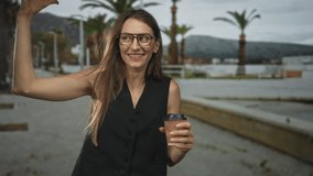 Young woman holding coffee cup and raising fist on seaside street promenade, bare arm visible; victory joy. - Powered by Shutterstock - Get 15% off with code: PIKWIZARD15