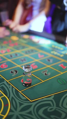 Casino table with roulette wheel in motion, with casino chips, tokens, the hand of croupier, dollar money and a group of gambling rich people playing bet on a party, blue poker table and deck of cards