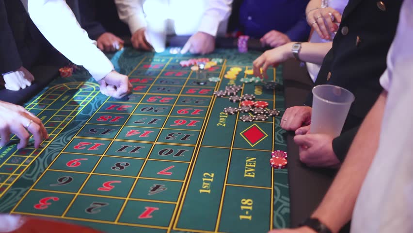 Casino table with roulette wheel in motion, with casino chips, tokens, the hand of croupier, dollar money and a group of gambling rich people playing bet on a party, blue poker table and deck of cards