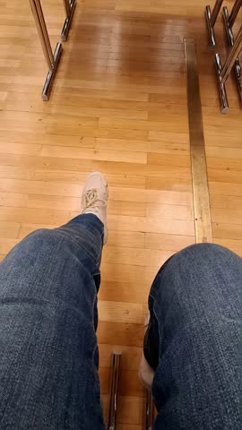 A man is seated at a table, playfully swinging his legs in the air. His casual demeanor creates a relaxed atmosphere in the indoor space. The wooden floor adds warmth to the scene.