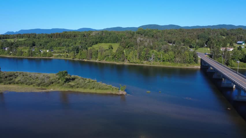 A black pickup truck crosses the Cascapedia River Bridge, a renowned salmon sport fishing river flowing from the Chic-Choc Mountains in the background under a cloudless blue sky, Quebec, Canada, 2025.