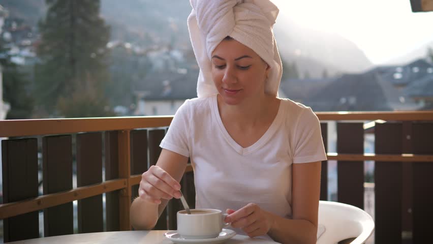 Beautiful woman with towel on head drinking cup of hot coffee on balcony in morning. Woman enjoying fresh air and amazing view of mountains at ski resort