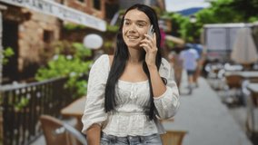 Woman holds smartphone to ear while smiling and talking on street cafe terrace; conversation leisure contentment. - Powered by Shutterstock - Get 15% off with code: PIKWIZARD15