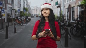 Woman in santa hat holds phone with both hands, pout expression on face in a narrow street wearing red top; holiday melancholy. - Powered by Shutterstock - Get 15% off with code: PIKWIZARD15