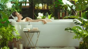 Woman with eyes closed finds serene relaxation in a bathtub, wearing headphones amidst lush plants and candles. Luxurious, tranquil scene epitomizes self-care and wellbeing in a home spa setting. - Powered by Shutterstock - Get 15% off with code: PIKWIZARD15
