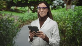 Woman scientist in lab coat wearing safety goggles taps tablet with fingers on park path; concentration field research. - Powered by Shutterstock - Get 15% off with code: PIKWIZARD15