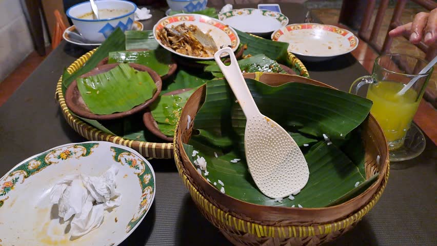Rainy atmosphere at a traditional Indonesian restaurant with joglo design, showing empty plates and bamboo platters with food remnants, surrounded by flies in a natural post-meal scene.